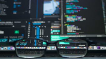 Glasses sitting on a desktop with large computer monitors showing code behind them.