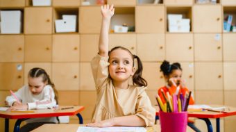 Three young girls in a classroom with one of them smiling and raising her hand.