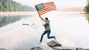 A man leaps from one rock to another in a river while carrying an American flag.