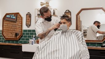 A young man with a protective mask over his nose and mouth gets a haircut from a man who is also wearing a protective mask.