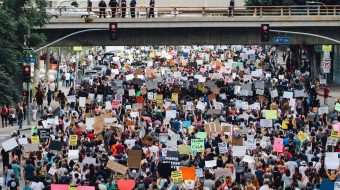 A crowd of protesters march on a city street, under a bridge.