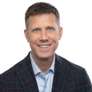 Brunette man in blue shirt and dark patterned jacket, smiling.